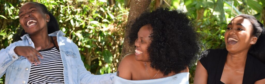 Three young African women, sat together, laughing outside.