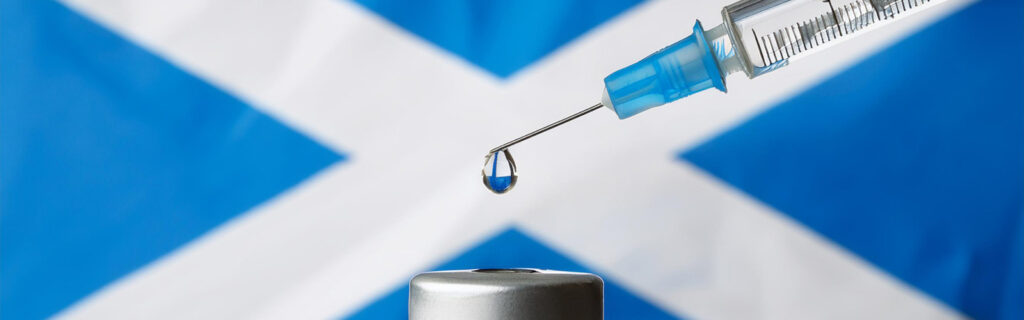 A needle drips a clear drop of fluid, in the background is the Scottish flag