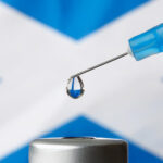 A needle drips a clear drop of fluid, in the background is the Scottish flag